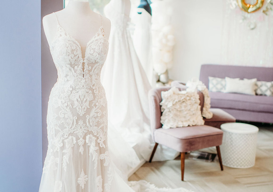 Sitting area in a bridal shop with a lavender couch, two patterned fabric side chairs, and a rack of bridal gowns